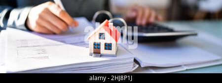 Mano della persona che fa il calcolo dell'imposta di proprietà usando il calcolatore del modello della casa Foto Stock