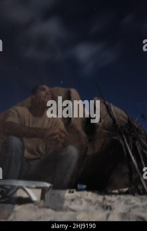 Foto notturna di un uomo seduto sulla spiaggia con una roccia di granito, illuminata dal chiaro di luna. Esposizione lunga e multipla Foto Stock