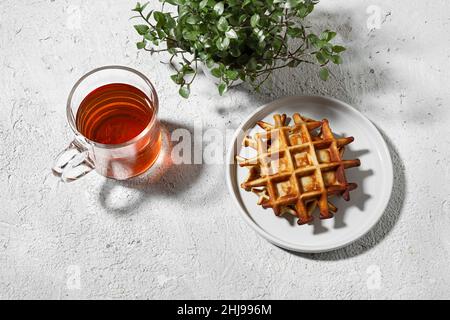Waffle belgi e una tazza di tè su un tavolo leggero con pianta di callisia, gustosa colazione concetto, vista dall'alto Foto Stock