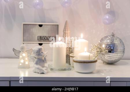 Il concetto di decorazione della casa per Natale. Candele accese, una ghirlanda, una palla da discoteca, un angelo figurine su una cassa bianca di cassetti. Luogo per Foto Stock