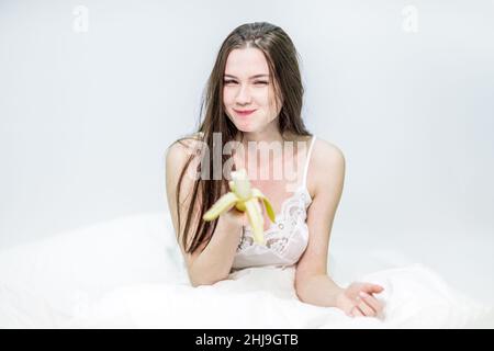 Una bella brunetta è fare colazione con frutta. La ragazza si siede sul letto, avvolta in una coperta, e ama mangiare una banana. Foto Stock