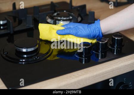 Stufa a gas di pulizia. Mano donna in guanto di gomma lucidando la superficie in vetro nero con panno giallo. Foto Stock