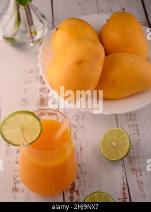 La foto mostra vetro trasparente con succo di mango fresco naturale su testurizzato vintage. Sullo sfondo, esotici frutti di mango gialli possono essere visti. In primo piano Foto Stock