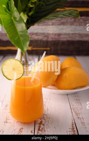 La foto mostra vetro trasparente con succo di mango naturale. Sullo sfondo, esotici frutti di mango gialli possono essere visti. In primo piano ci sono fette di agrumi Foto Stock