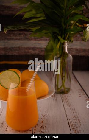 La foto mostra vetro trasparente con succo di mango naturale. Sullo sfondo, esotici frutti di mango gialli possono essere visti. In primo piano ci sono fette di agrumi Foto Stock