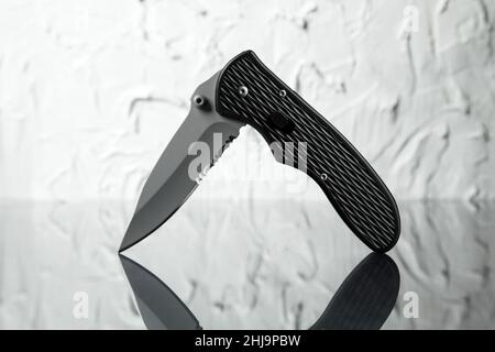 Fotografia di prodotto di un moderno ed elegante coltello nero tascabile in posizione semi aperta su una superficie in vetro riflettente. Foto Stock