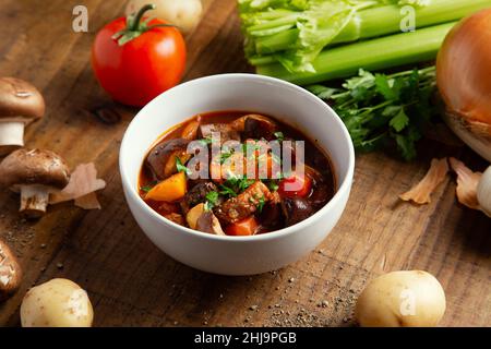 Accogliente ciotola per cena con stufato di manzo invernale con patate fresche crude, sedano, cipolle, pomodori e funghi su un rustico tavolo in legno. Foto Stock