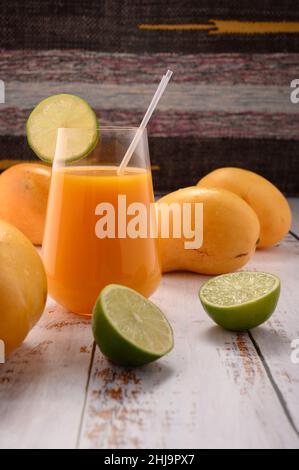 La foto mostra vetro trasparente con succo naturale di mandarino fresco su testurizzato vintage. Sullo sfondo, esotici frutti di mango gialli possono essere visti. In rinuncia Foto Stock