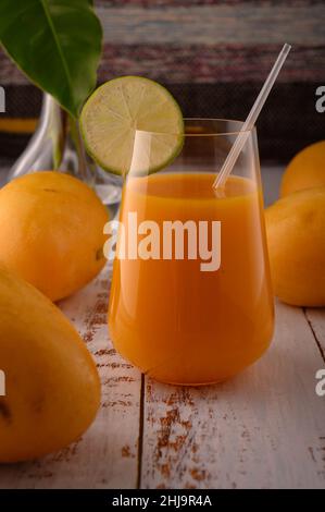 La foto mostra vetro trasparente con succo di mango fresco naturale su testurizzato vintage. Sullo sfondo, esotici frutti di mango gialli possono essere visti. In primo piano Foto Stock