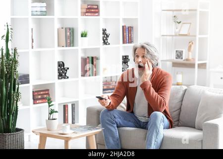 Uomo anziano scioccato che guarda la TV sul divano di casa Foto Stock