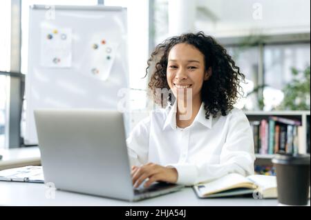 Bella ragazza afroamericana positiva, con capelli ricci, direttore vendite, agente immobiliare, segretario, si siede in un ambiente di lavoro in un ufficio moderno, lavora in un notebook, guarda la fotocamera, sorridi amichevole Foto Stock
