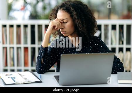 Mal di testa, affaticamento. Esausta, strana, strana donna afroamericana, lavoratore d'ufficio, manager, si siede sul posto di lavoro in ufficio, chiuso gli occhi, massaggia il naso del ponte, ha bisogno di riposo, rottura Foto Stock
