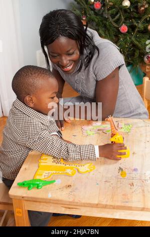 ragazzo di 2 anni che gioca con giocattoli giraffa di diverse dimensioni con puzzle di cartone giraffa davanti a lui, incoraggiato dalla madre Foto Stock