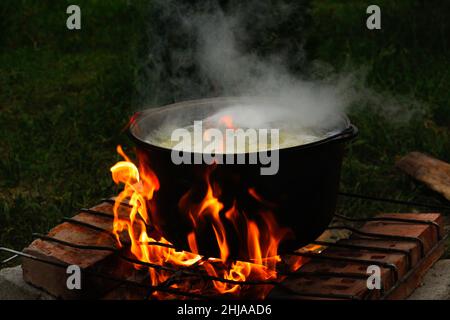 Cottura della zuppa di pesce nel bowler di ferro sopra un falò. La zuppa di pesce bolle in calderone al palo. Zuppa in una pentola nel fuoco. Campeggio di notte. Verde natura Foto Stock