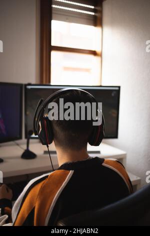 Vista posteriore di concentrato di giovane giocatore in cuffie e occhiali che usano il computer per la riproduzione di gioco a casa Foto Stock