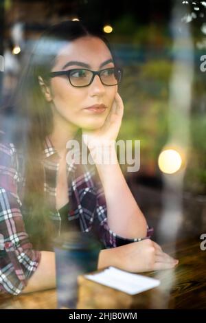 Annoiato o ragazza pensiva guardando via nella finestra con telefono sul tavolo presso il caffè università indossando occhiali. Giovane studentessa in casual guardando sognato fuori dalla finestra del caffè di navigazione dei social media. Foto Stock