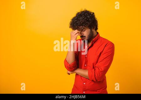 Problema, depressione o malattia. Ritratto di solo triste bel giovane barbuto uomo in camicia in piedi, tenendo la testa giù e piangendo. Studio interno girato isolato su sfondo arancione Foto Stock