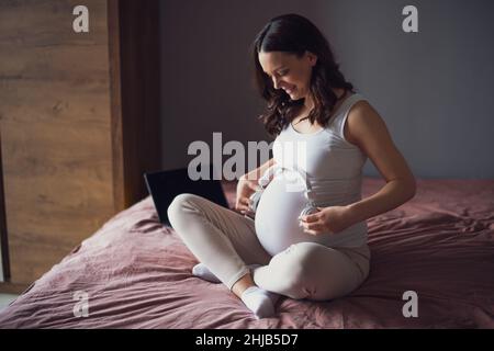 Donna incinta felice rilassarsi a casa. È seduta sul letto in camera da letto e suona la musica al suo bambino nello stomaco. Foto Stock