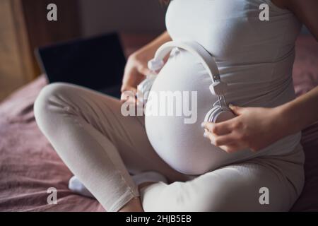 Primo piano di una donna incinta che si rilassa a casa. È seduta sul letto in camera da letto e suona la musica al suo bambino nello stomaco. Foto Stock