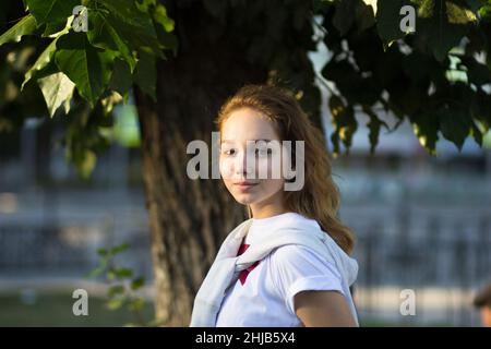 Ritratto della giovane donna caucasica nel parco estivo guardando la macchina fotografica in piedi vicino all'albero Foto Stock