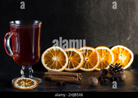 Bicchiere di VIN brulé, spezie e pezzi di arance su sfondo nero Foto Stock