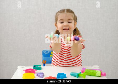 Piccola bambina sorridente in T-shirt a righe gioca con plastilina colorata al tavolo bianco, gioca l'impasto a casa Foto Stock