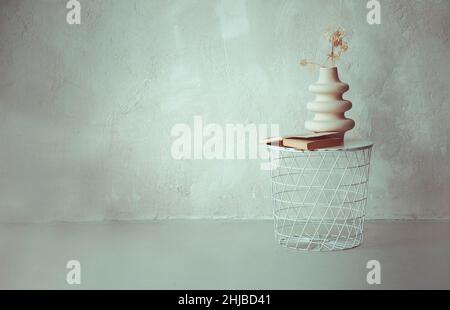 Libro e asciugato ramoscello in vaso di ceramica su bianco metallo rotondo comodino contro la parete di calcestruzzo beige chiaro in stile loft interno semplice appartamento, copia Foto Stock