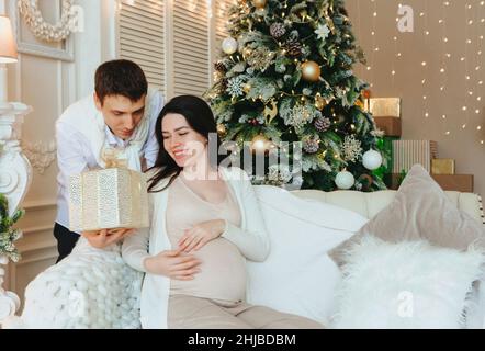 Giovane marito amorevole che dà la vacanza presente a felice sorpresa moglie incinta mentre festeggia il Natale o Capodanno insieme. Futuro della famiglia giovane par Foto Stock