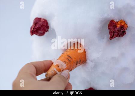 Primo piano di mano facendo nevaio mettere carota come naso sulla faccia con occhi rossi sul cortile di giorno mentre si cammina. I genitori che trascorrono il tempo con Foto Stock