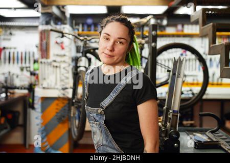 Meccanico femminile con serrature verdi che guardano la telecamera mentre si trova vicino al banco di lavoro in un negozio di riparazione professionale. Foto Stock