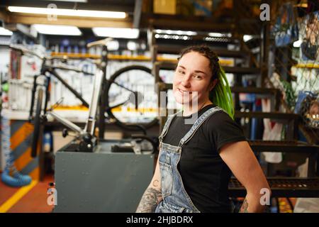 Una donna felice con lucchetti luminosi sorridenti e guardando la telecamera mentre si trova vicino ai gradini durante il lavoro in un negozio di riparazione biciclette. Foto Stock