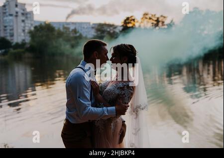 Sposarsi e sposarsi con le bombe fumogene di colore blu e giallo nel parco estivo. La sposa e lo sposo con il fumo colorato sullo sfondo di Foto Stock