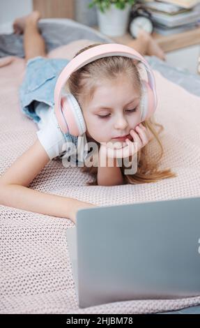 Sorridente bambina felice utilizzando il laptop in cuffie wireless a letto a casa. Foto Stock