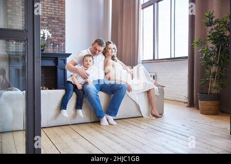 Famiglia caucasica con un bambino piccolo e una moglie incinta con un ventre in un soggiorno con pavimento in legno. Foto Stock