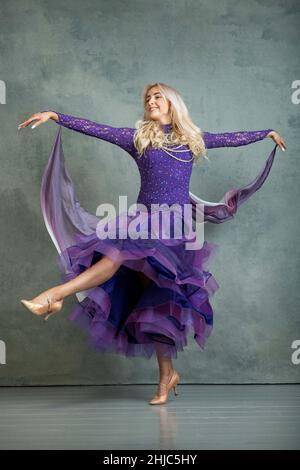 Ballerina Bionda femmina in movimento di danza scorrevole in abito da ballo viola, sullo sfondo grigio dello studio fotografico Foto Stock