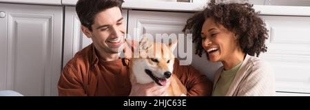 felice coppia interrazziale sorridendo vicino divertente shiba inu cane in cucina, banner Foto Stock