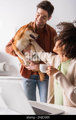 donna afroamericana con tazza di caffè che accarezzava il cane shiba inu nelle mani di un ragazzo sorridente Foto Stock