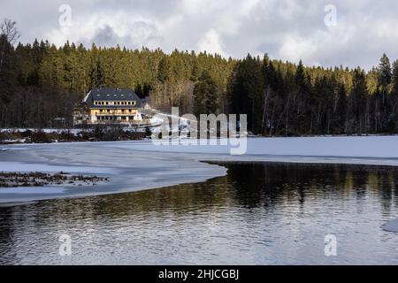 Lenzkirch, Germania. 28th Jan 2022. Uno strato di ghiaccio galleggia sulla Windgfällweiher vicino a Schluchsee, mentre sullo sfondo si possono vedere conifere e una casa della Foresta Nera. Credit: Rin/dpa/Alamy Live News Foto Stock