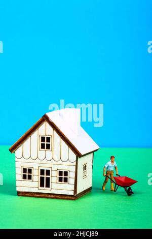 Immagine concettuale di un uomo in miniatura con una carriola accanto ad una casa modello Foto Stock