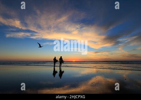 I surfisti su una spiaggia lucida con bassa marea guardano un colorato tramonto invernale dopo il surf a Torrey Pines, San Diego, California. Foto Stock