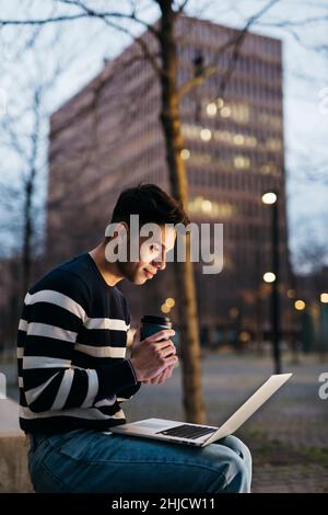 Giovane uomo seduto su panca, con un computer portatile e un caffè da bere Foto Stock