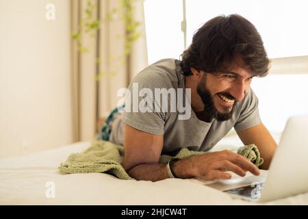 Giovane uomo che lavora a casa dalla sua camera da letto con un computer portatile sul suo letto. Uomo sdraiato. Uomo sdraiato. Notebook grigio per il lavoro. Accogliente e confortevole. Ho Foto Stock