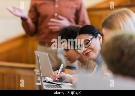 Sorridente studente asiatico iscritto su notebook vicino a amici multietnici e notebook in università Foto Stock
