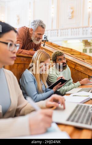 Insegnante sorridente che guarda gli studenti interrazziali con notebook e notebook in università Foto Stock