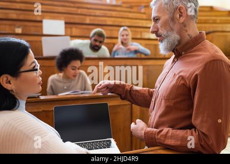 Insegnante maturo che punta al laptop con schermo vuoto vicino a studente asiatico in università Foto Stock