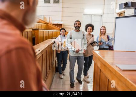 Allegri studenti multiculturali con notebook e dispositivi in piedi vicino a un insegnante sfocato in università Foto Stock