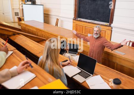 Insegnante sorridente che tiene gesso vicino a studenti multietnici in auditorium universitario Foto Stock