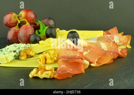 Vassoio con prosciutto di Schwarzwald, formaggi, noci, olive nere. Decorato con basilico. Cibo sul backgroud ardesia. Spazio di copia Foto Stock