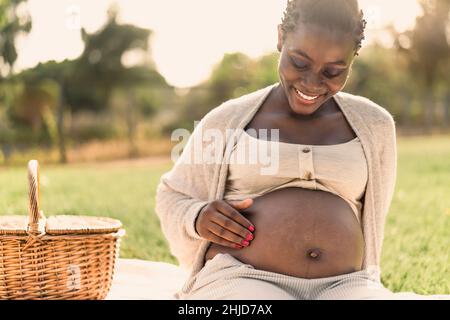 Donna africana carezzando il suo ventre incinta mentre fa un picnic nel parco - concetto di stile di vita di maternità Foto Stock