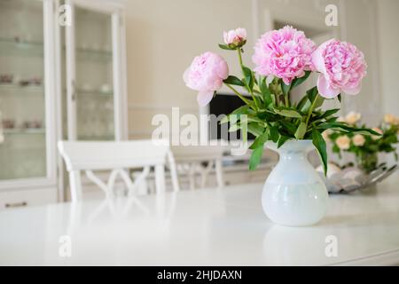 Fiori di peonia rosa in fiore con foglie verdi e lussureggianti messi in vaso su tavola bianca in camera luminosa con mobili moderni a casa primo piano Foto Stock